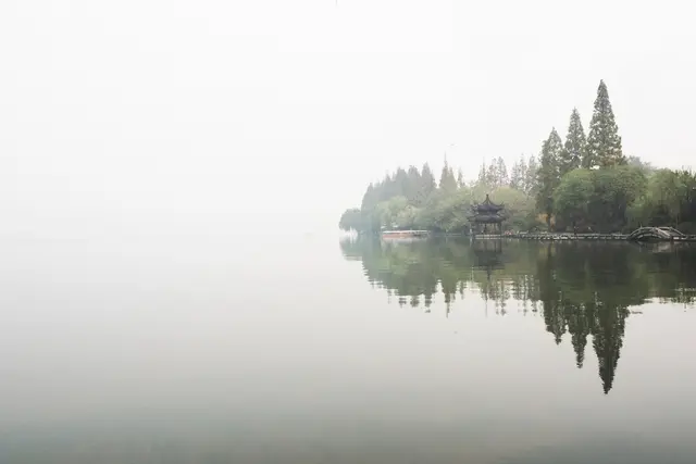 Een mistig meer met een traditioneel paviljoen tussen de bomen, alles weerspiegeld in het kalme water, wat een serene scène creëert.